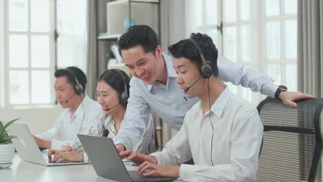 a man coming to help one man of three asian call center agents in headsets to work with computer while two of his colleagues are speaking with customer at the office