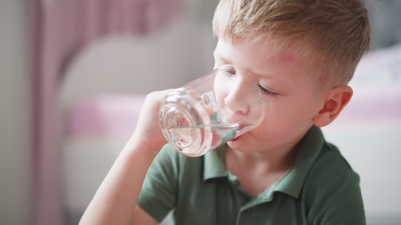 Close up of young boy wearing green shirt drinking water from transparent glass indoors with soft light and blurred pink and white background