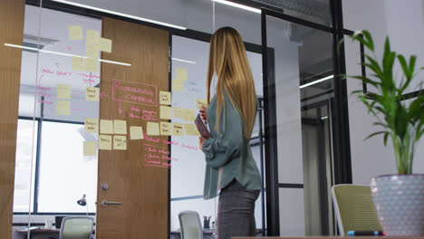 caucasian businesswoman holding tablet brainstorming using memo notes on glass wall in office