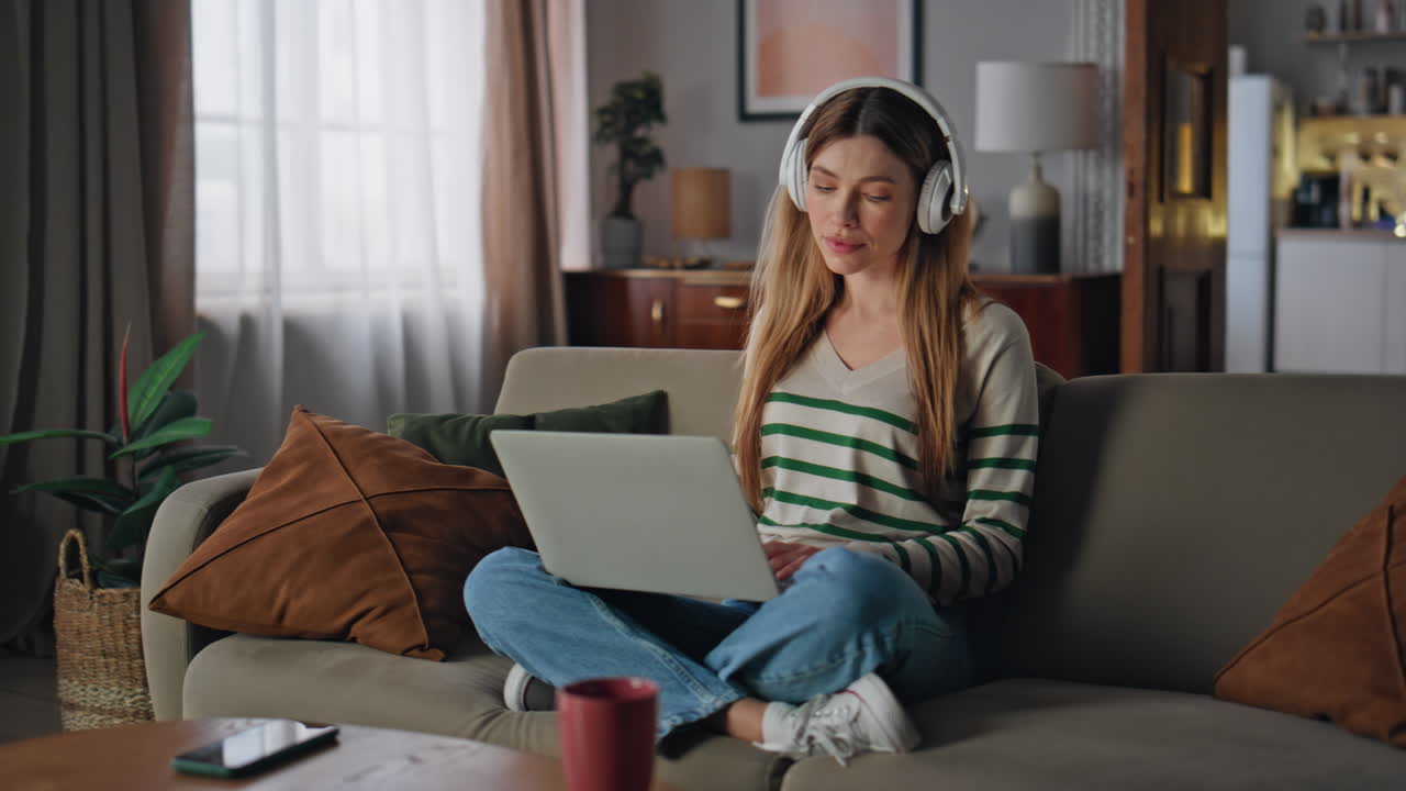 Ragazza spensierata che naviga sul laptop ascoltando musica con cuffie wireless sul divano.