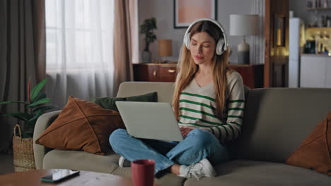 Carefree girl surfing laptop listening music in wireless headphones at couch.