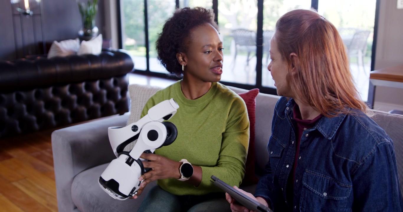 Diverse female friends examining helmet after woman lifting it, using tablet for feedback on sofa