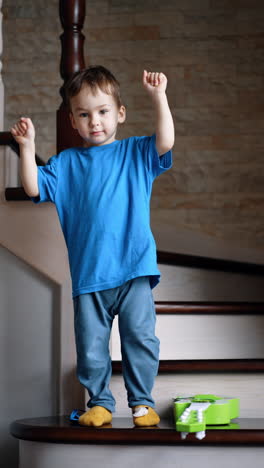 dark-haired toddler boy wearing blue t-shirt stands on the stairs. kid waves his hands and then goes down looking at camera. vertical video.