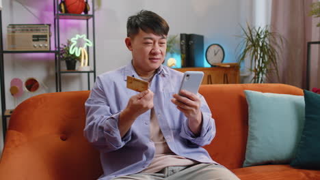 asian adult man using credit bank card and smartphone, transferring money, purchases online shopping