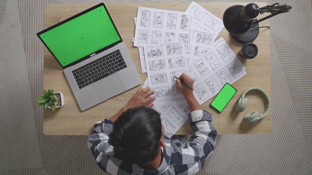 vista superior de un artista masculino asiático dibujando un guión gráfico para la película en la mesa con un teléfono inteligente de pantalla verde y una computadora portátil en el estudio