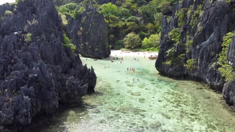 Wide-low-Aerial-Push-in-View-towards-Hidden-Beach-and-its-jungle-surrounded-white-sand-and-karst-Rock-Cliffs,-Tourists-swimming-in-tropical-shallow-turquoise-water-Lagoon,-Tour-C-El-Nido,-Philippines