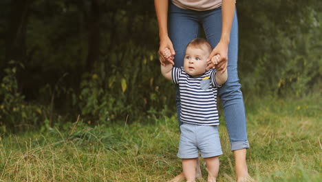 portrait of a baby boy taking his first steps in the grass while holding his mother's hands