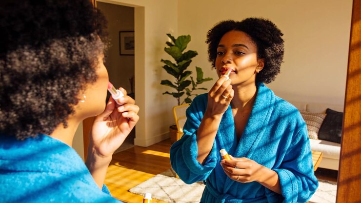 Raising lip product, Black woman wearing blue robe applying lipstick at mirror, preparing day