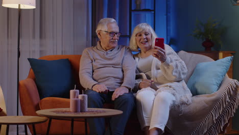 Sonriente-Pareja-De-Ancianos-Abuela-Abuelo-Haciendo-Videollamadas-En-Línea-En-El-Teléfono-Móvil-En-El-Sofá-De-Casa