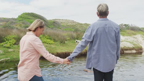 Smiling-senior-caucasian-couple-holding-hands-at-river-with-dog-outdoors