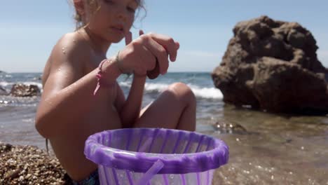 young european girl picking up stones and putting it into purple bucket at the beach in spain