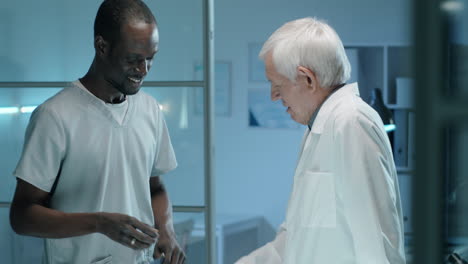 Joven-Trabajador-De-Laboratorio-Negro-Dándose-La-Mano-Y-Hablando-Con-Un-Científico-Senior