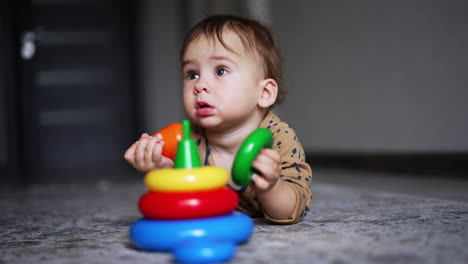 lovely boy lies on carpet indoors with a toy in front of him. cute kid gathering the parts of bright toy pyramid. blurred backdrop.