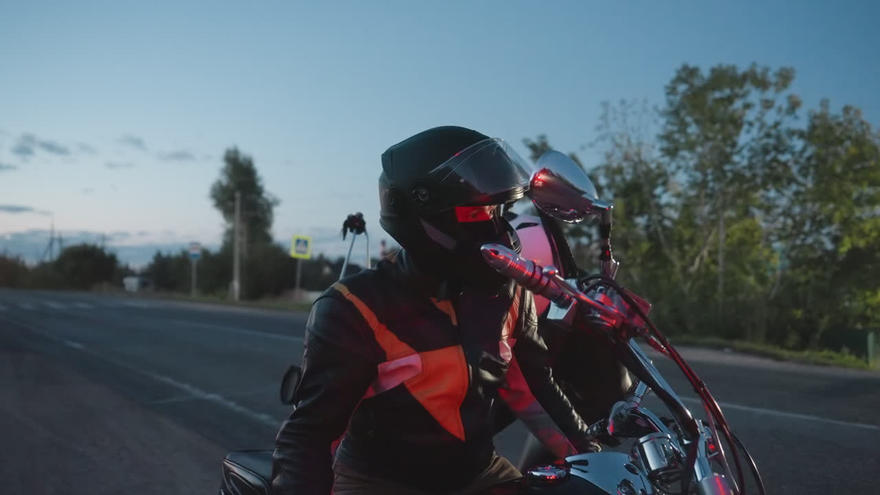 Motorcycle riders in helmets and leather jackets climb onto chrome motorbike on roadside at dusk, with red sunset glow and blurred headlights in distance creating energetic evening travel moment