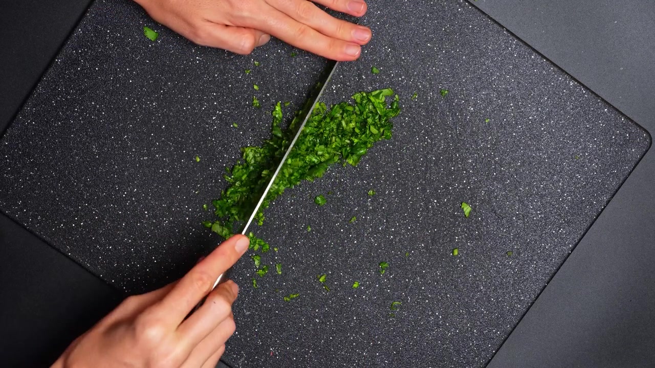 Person mincing fresh green cilantro on dark board. Top view of hands using a knife to finely chop green coriander herbs on a black cutting board.