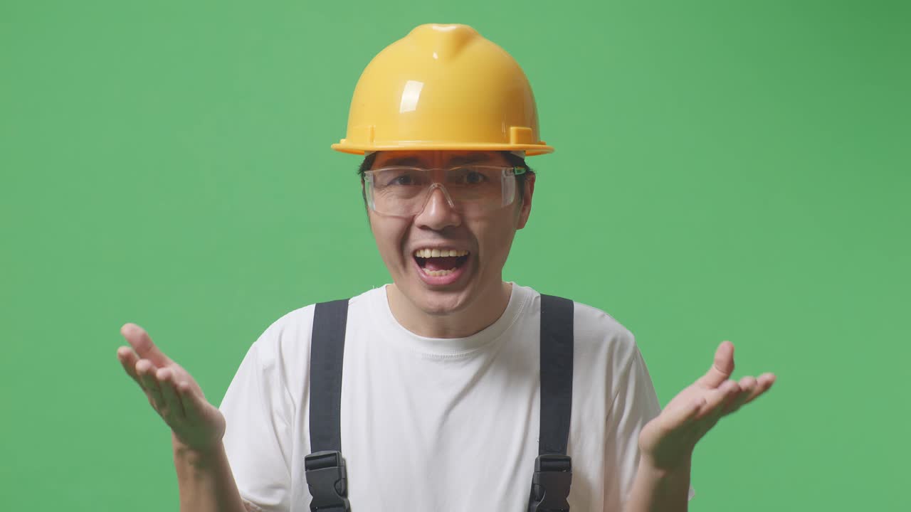 동양인 노동자가 보호 안경과 안전 헬을 착용하여 녹색 스크린 배경 스튜디오에서 의심스럽게 서 있습니다.