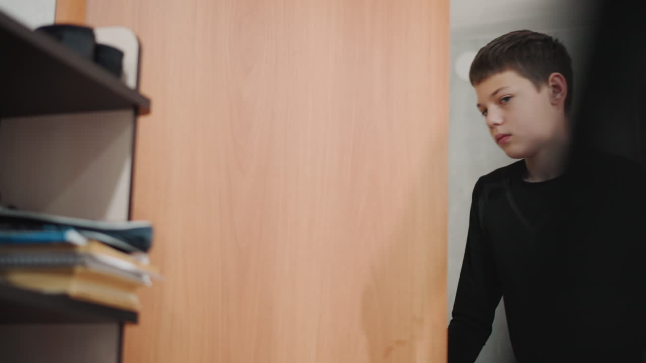 Il nipote adolescente bianco entra in una piccola area studio con scrivania e scaffali, libri impilati. Un movimento discreto mentre passa davanti all'inquadratura dirigendosi verso la postazione di lavoro, in un'atmosfera di apprendimento domestica e tranquilla.
