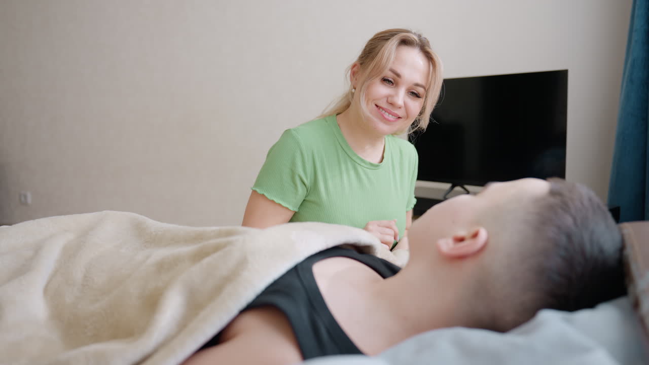 Madre premurosa accanto al figlio sotto le coperte che lo guarda con affetto mentre si sveglia nella camera da letto con il televisore sullo sfondo che mostra l'amore mattutino di una famiglia