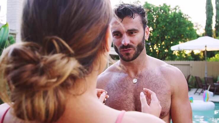Romantic Hispanic Couple With Woman Pushing Man Into Swimming Pool At Outdoor Summer Pool Party