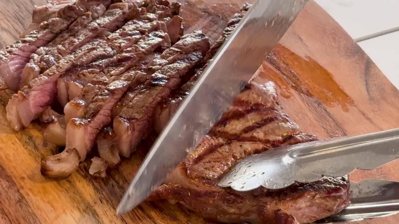 A juicy, grilled wagyu beef steak is sliced with a chef’s knife and tongs on a wooden cutting board under bright, natural lighting in a kitchen setting