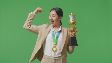 asian business woman in a suit with a gold medal and trophy flexing her bicep and smiling to camera on green screen background in the studio