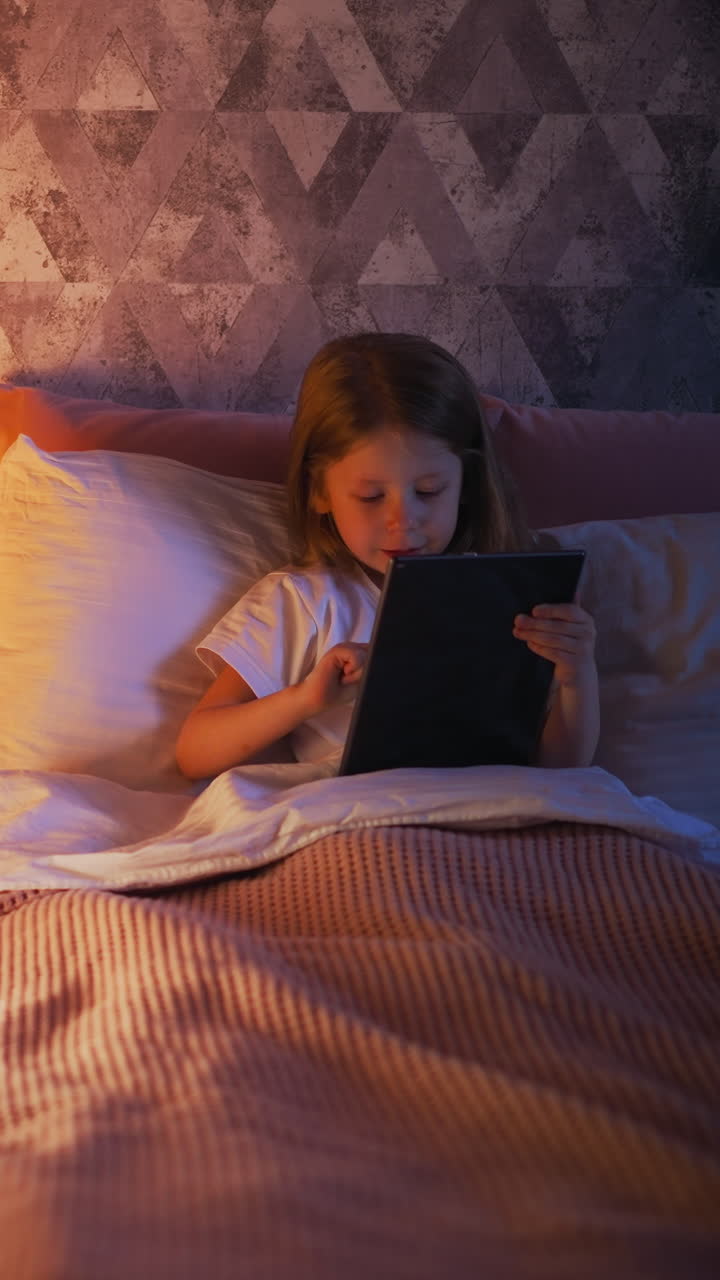 Happy little girl plays on contemporary tablet in comfortable bed with pillows and warm bedspread near children tent at lamp light and corner shadows slow motion