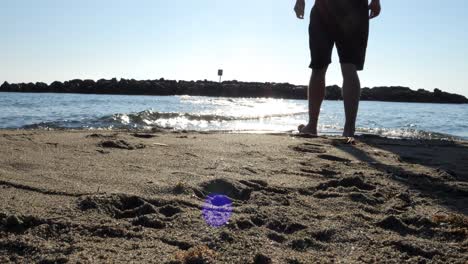 Low-angle-static-shot-in-front-of-the-beautiful-beach-in-Le-Barcarès-in-France-with-view-of-the-calm-waves-and-stones-in-the-water-and-a-vacationer-crossing-the-water-on-vacation