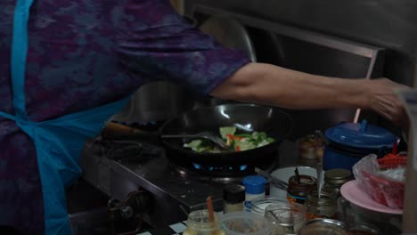 a women wearing a blue apron frying vegetables in a wok next to a table full of ingredients in a street kitchen, bangkok - thailand