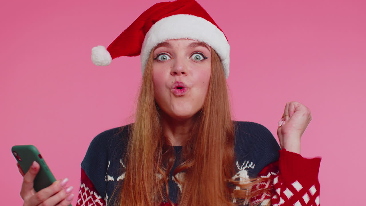 mujer con elegante suéter navideño mirando la pantalla del teléfono inteligente regocijándose sinceramente por ganar éxito y suerte