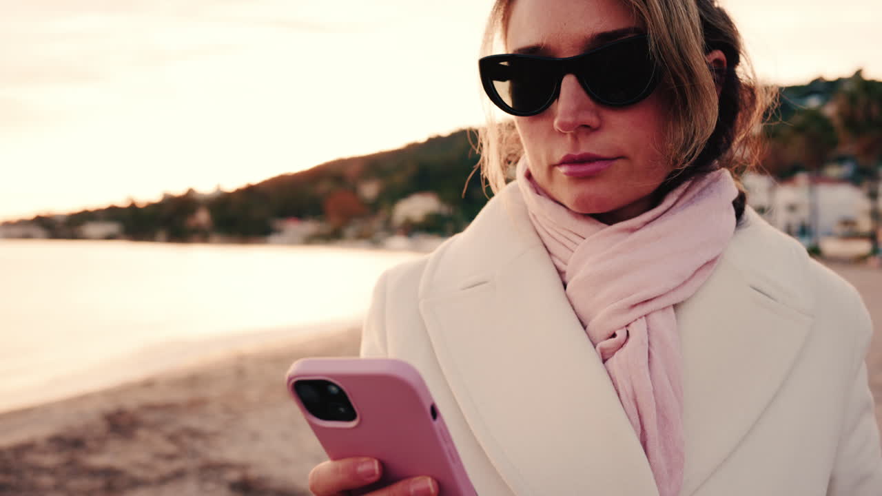 Donna elegante con cappotto bianco, sciarpa e occhiali da sole che controlla il telefono mentre è in piedi su una spiaggia al tramonto