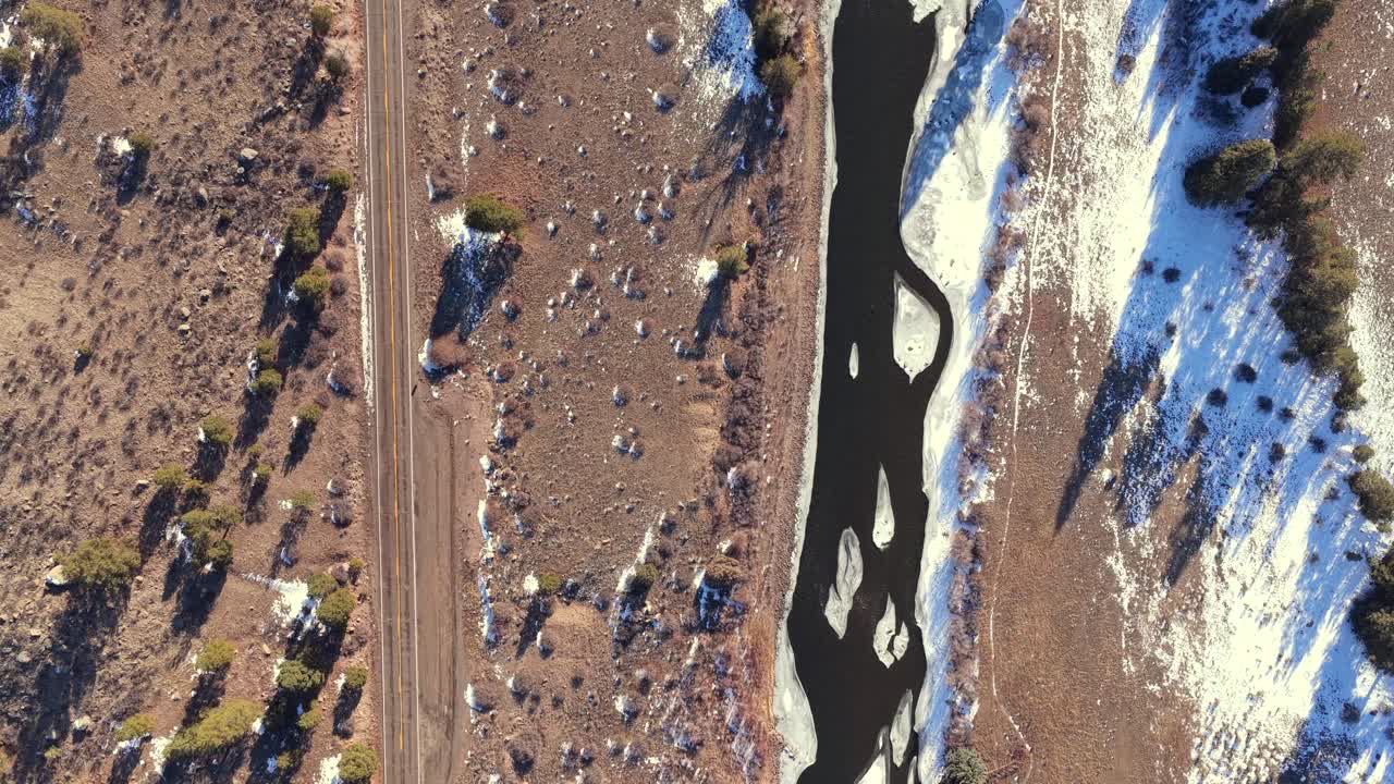 A cinematic top-down aerial shot follows a paved highway running parallel to a partially frozen river. This snowy winter landscape is perfect for automotive commercials and travel brand storytelling