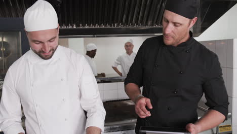 focused caucasian male chef instructing trainee male chef using tablet in kitchen, slow motion