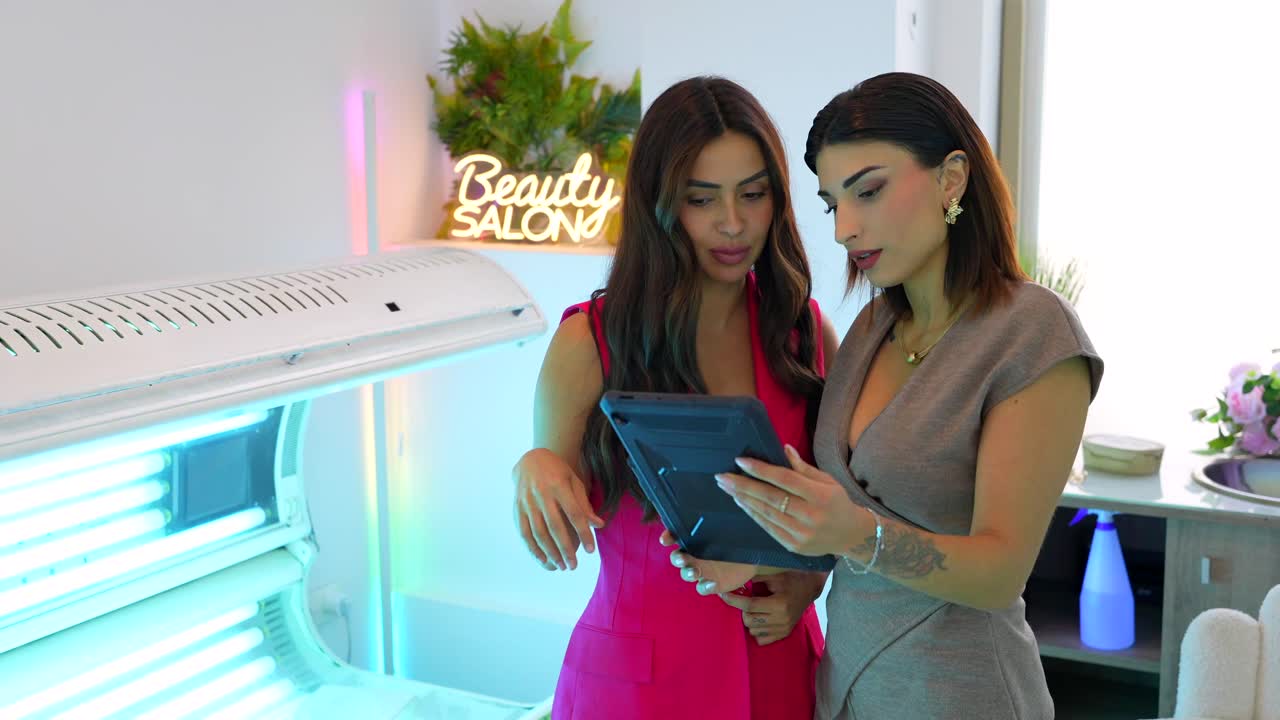 Two women discussing something on a tablet in a beauty salon near a tanning bed
