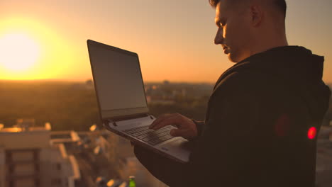 hacker using laptop on rooftop with city view and forex chart. hacking and stats concept. a man at sunset in slow motion writing software code on a laptop