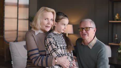 happy grandparents with their little granddaughter smiling and waving at camera in a modern living room at home