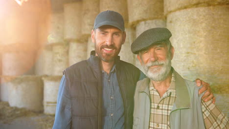 caucasian old gray-haired man and young man standing outside at stable with hay stocks and looking at camera