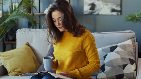 Young-Good-Looking-Lady-Flipping-Pages-And-Reading-A-Fashion-Magazine-While-Drinking-Hot-Tea-And-Coffee-And-Sitting-On-The-Couch-In-The-Living-Room