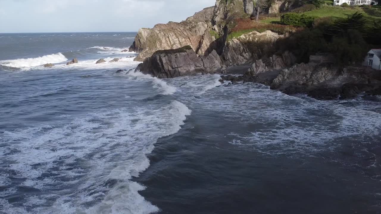 Aerial drone tracking shot following a wave from a rocky cliff coastline to an old mill house on the shore - Lee Bay, Beach, Ilfracombe, Devon, England