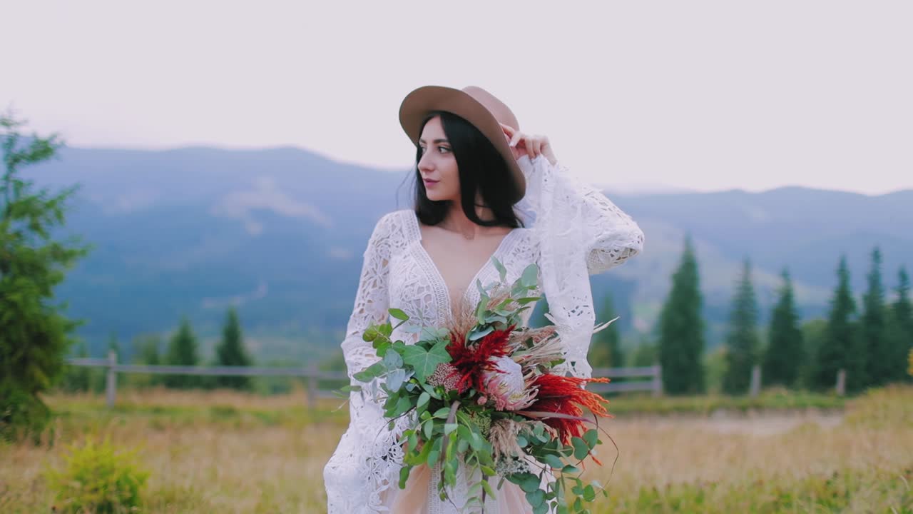 Half-length portrait of a bride in hat outdoors. Beautiful mysterious woman in white dress holds bouquet of flowers on the mountains background.