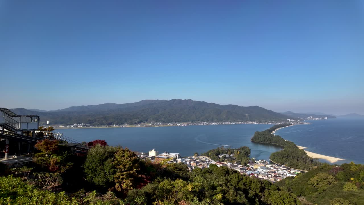 Scenic view of Amanohashidate Sandbar, a three kilometer isthmus, gracefully stretching across Miyazu Bay, framed by lush greenery and a vibrant blue sky. Establishing shot