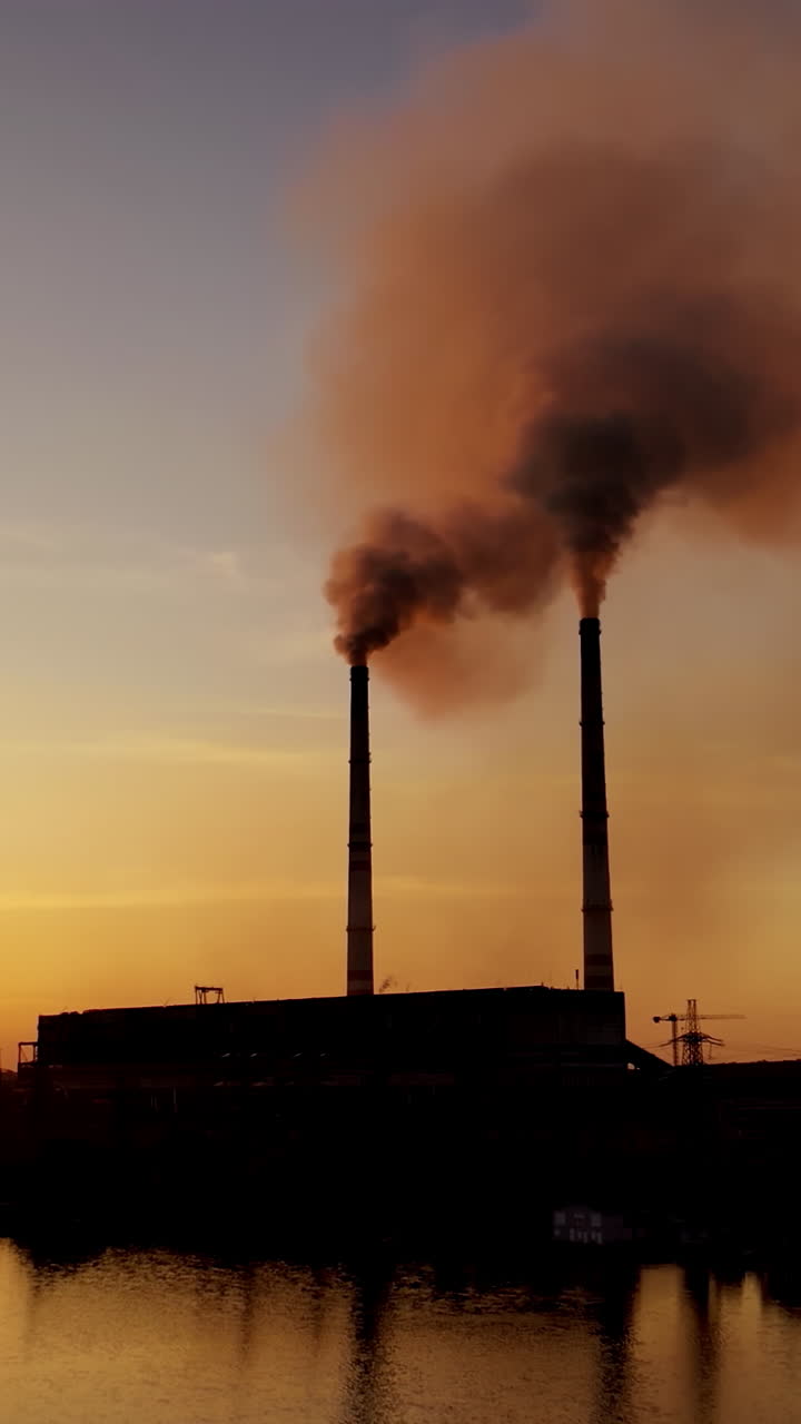 Industrial factory near the river at sunset. Smoke comes out of the pipes of a huge plant in the evening. Environmental pollution. Vertical video