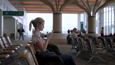 Una-Joven-Está-Sentada-En-La-Sala-Del-Aeropuerto-Y-Mira-Su-Teléfono-Celular