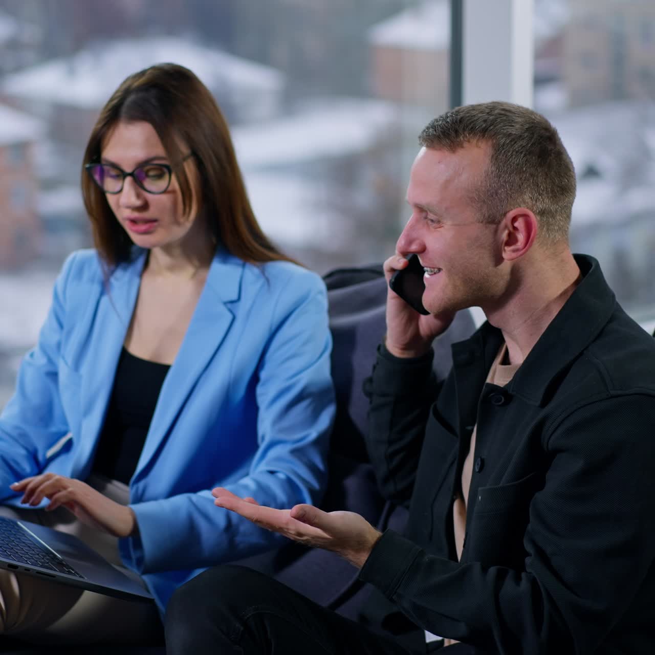 Company employees sit in bean bag chairs looking at the laptop in woman's hands. Man is holding his smartphone at the ear but points at computer with his hand