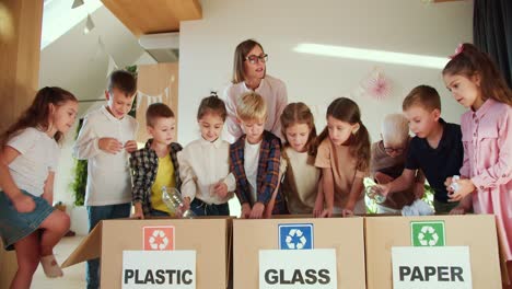 children, at the command of a teacher, women in a white shirt, put garbage into the right boxes separately, plastic, glass and paper in their club for preparing children for school. the woman teaches children how to properly dispose of trash