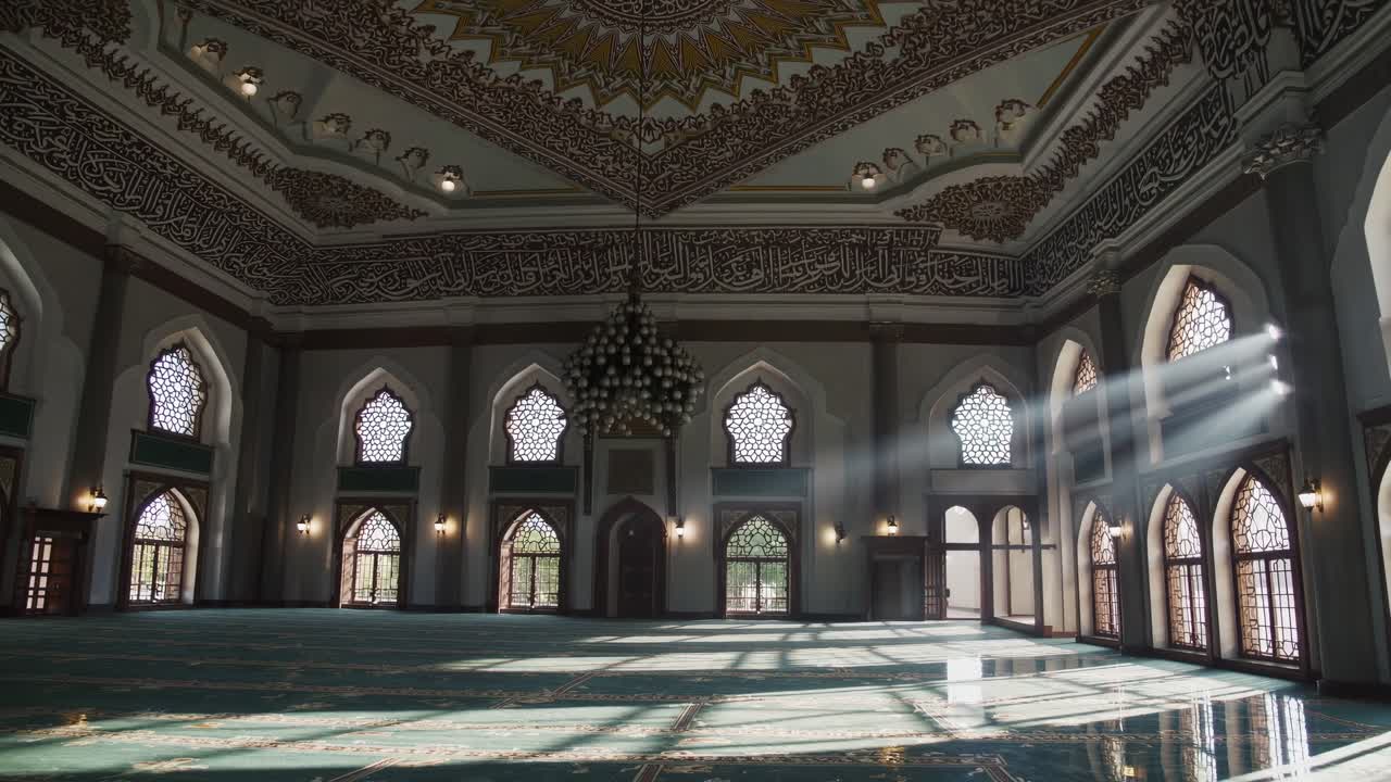 Sunbeams are streaming through arched windows into a large, empty mosque interior, illuminating the ornate ceiling, walls, and patterned carpet, creating a serene and spiritual atmosphere