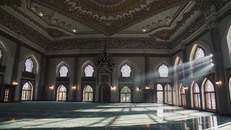 sunbeams are streaming through arched windows into a large, empty mosque interior, illuminating the ornate ceiling, walls, and patterned carpet, creating a serene and spiritual atmosphere