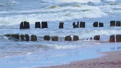 Olas-Tormentosas-Rompiendo-Contra-El-Viejo-Muelle-De-Madera-En-La-Playa,-Día-De-Primavera-Nublado,-Mar-Báltico,-Letonia,-Pape,-Primer-Plano-Medio-Desde-La-Distancia