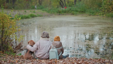 woman wraps arm around dog and child while sitting on blanket of fallen leaves beside calm lake, then slowly lowers hand in gentle gesture of affection as blurry people walk in background distance