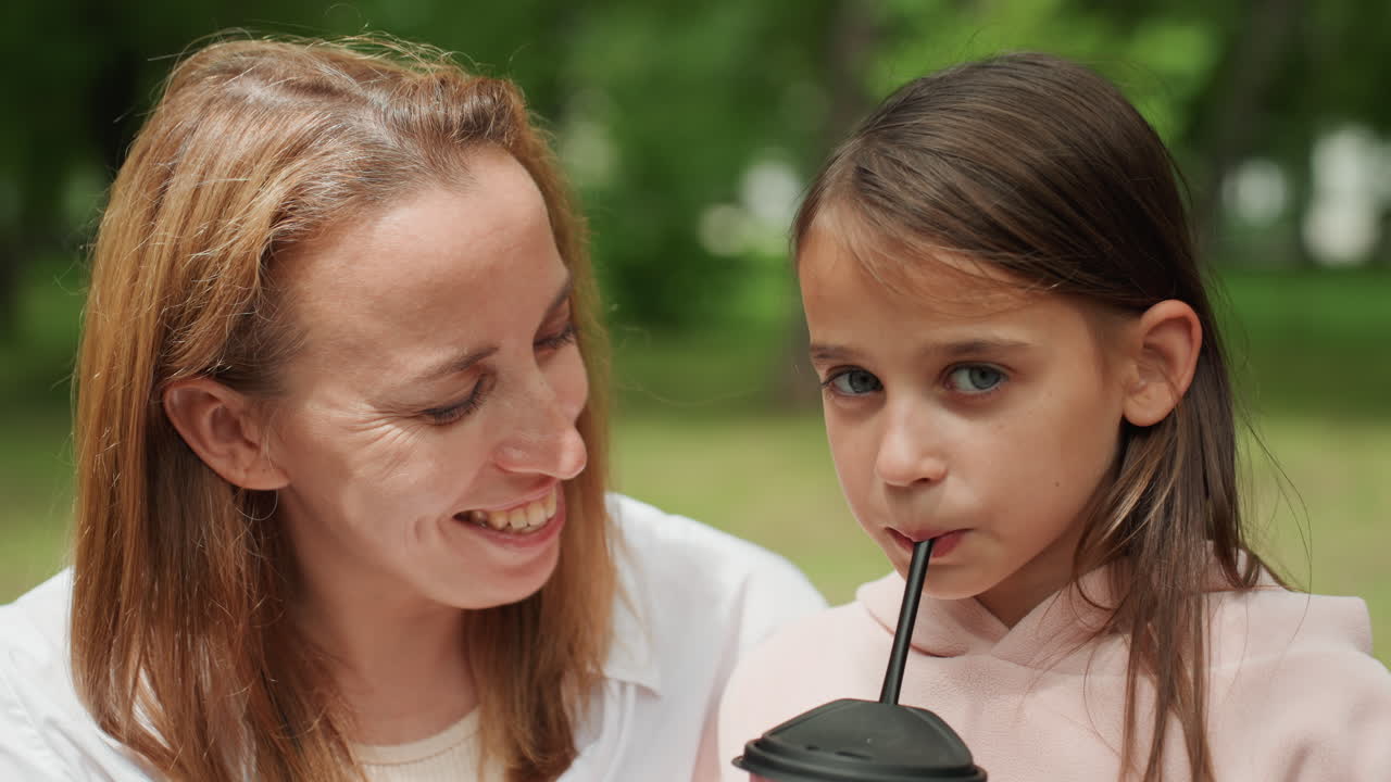 Seduti nel parco a condividere una bevanda fresca. Bambini e madre si connettono durante la condivisione di una bevanda all'aperto sotto il sole. Madre e figlia si scambiano sorrisi sorseggiando una bevanda rinfrescante in un lussureggiante ambiente del parco.