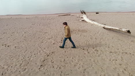 Aerial-drone-shot-of-a-young-man-walking-over-a-sand-beach,-log-laying-on-beach-and-sea-in-background